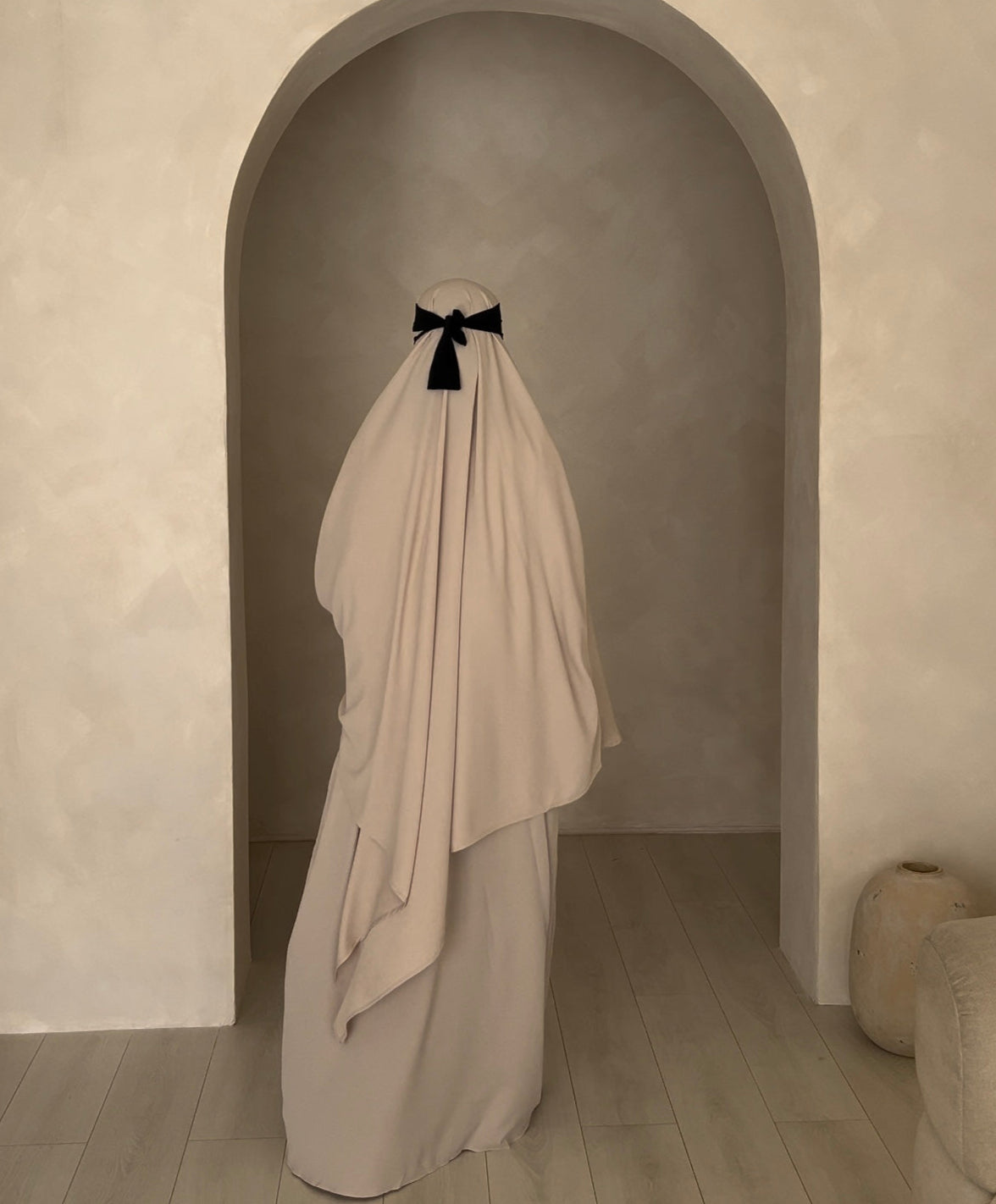 Person wearing a long, light-colored dress with a black bow in a room with an archway.